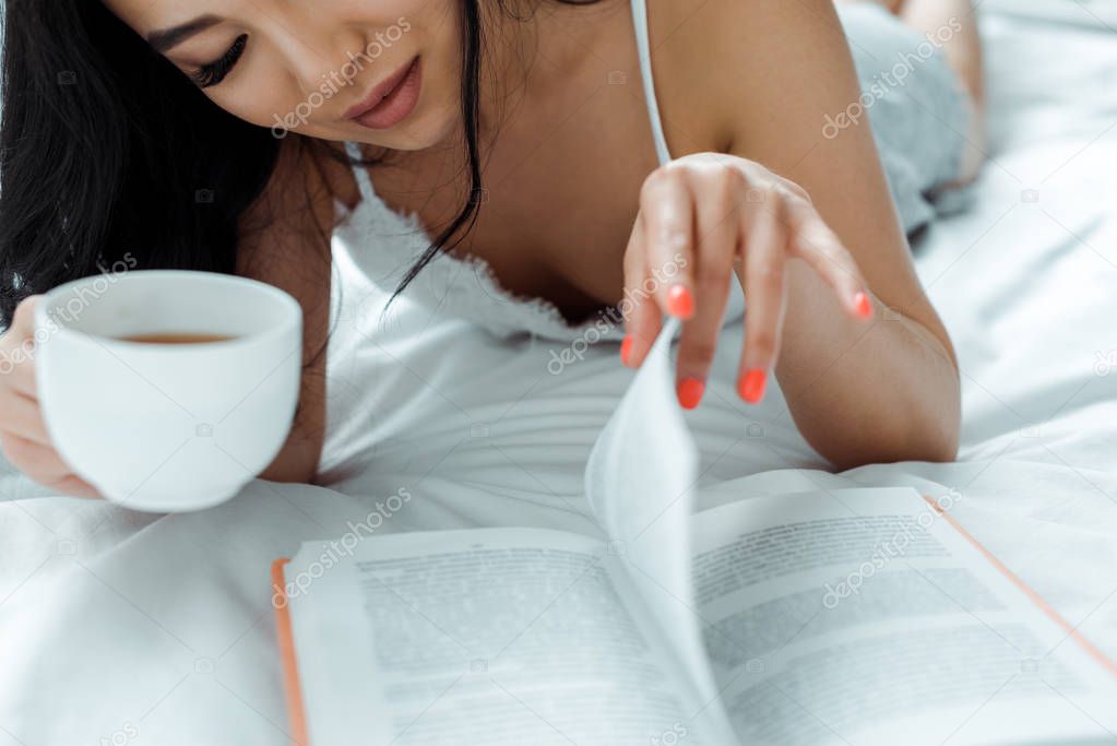 cropped view of asian woman lying on bed, reading book and holding cup of tea 