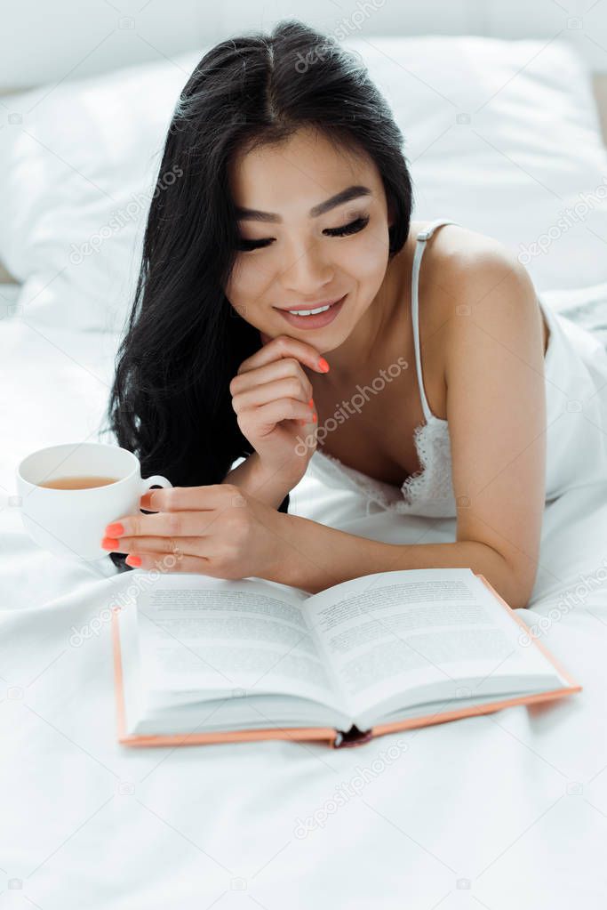 selective focus of happy brunette asian woman reading book and holding cup of tea 