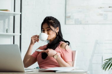 selective focus of attractive asian woman with closed eyes holding saucer and drinking tea  clipart