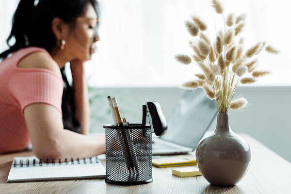 selective focus of vase with willow branches and pen holder near asian woman 
