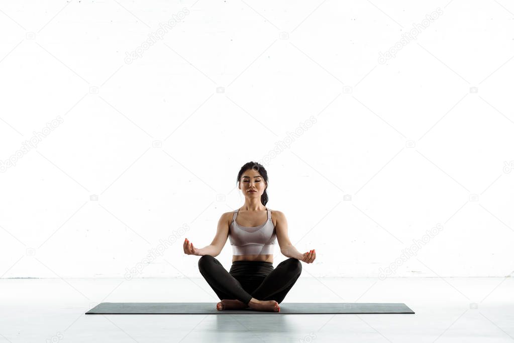 calm asian woman sitting on yoga mat and doing exercise on white