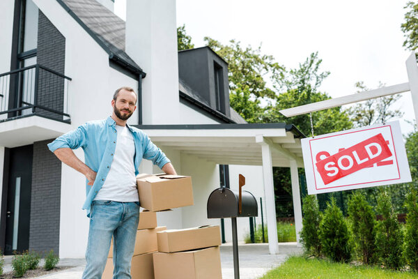 handsome man standing with hand on hip and holding box near house and board with sold letters 