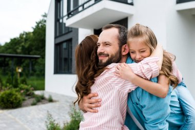 cheerful bearded man and happy kid hugging with woman near house  clipart
