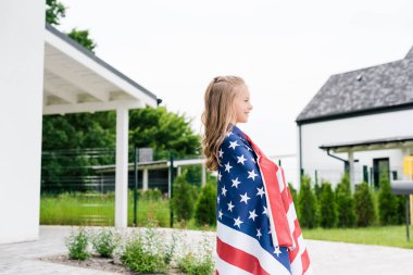 evin yakınında Amerikan bayrağı ile dururken gülümseyen mutlu çocuk yan görünümü 