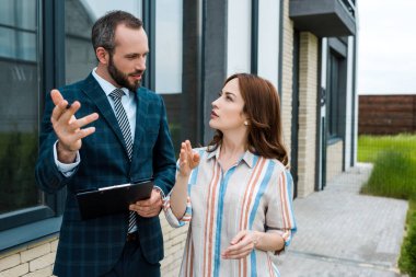 handsome bearded broker holding clipboard and gesturing near woman  clipart