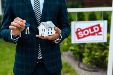 cropped view of realtor holding carton house model and keys near board with sold letters  clipart