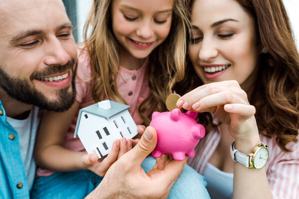 selective focus of happy woman putting golden coin in piggy bank near husband and daughter 