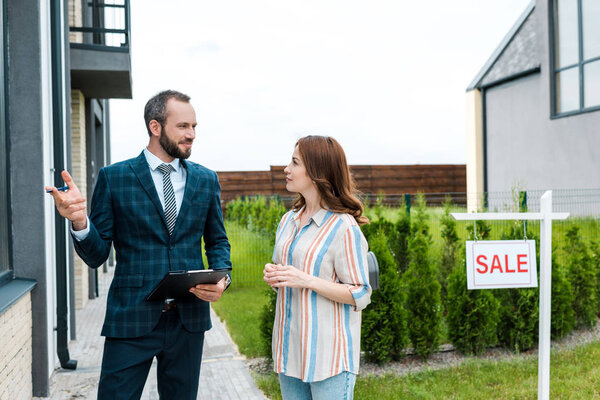 handsome bearded broker holding clipboard and gesturing near attractive woman and board with sale letters 