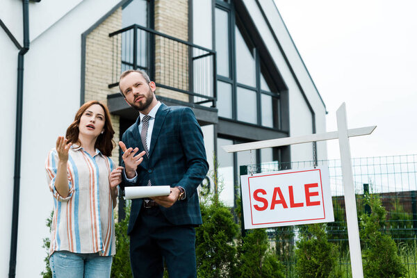 handsome broker holding clipboard and gesturing near attractive woman and board with sale letters 