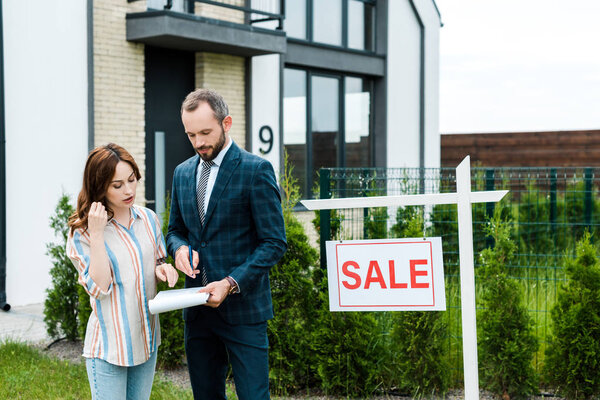 attractive woman looking at clipboard while standing near broker and house 