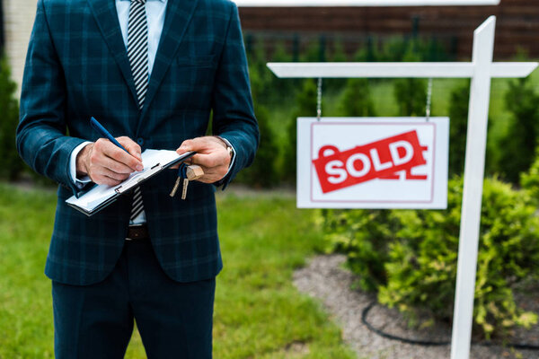 cropped view of broker writing while holding clipboard near board with sold letters 