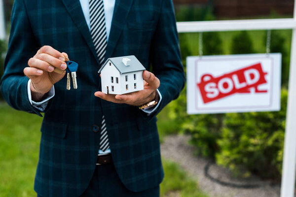 cropped view of realtor holding carton house model and keys near board with sold letters 