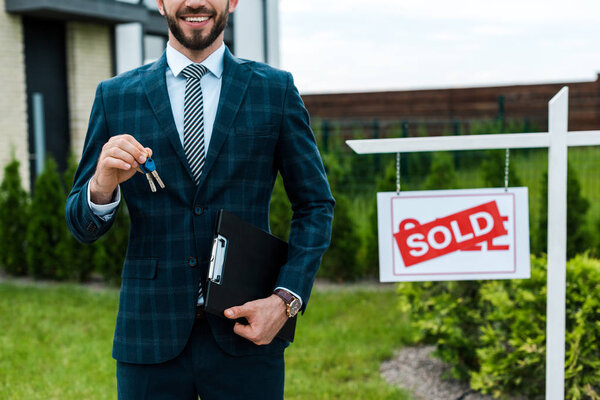 cropped view of cheerful broker holding keys and clipboard near board with sold letters 