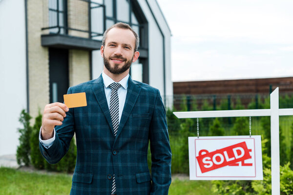 happy bearded man in suit holding credit card near house 