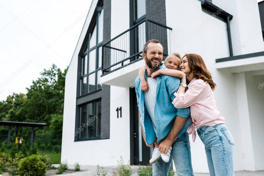 happy bearded man piggybacking daughter near cheerful wife and home 