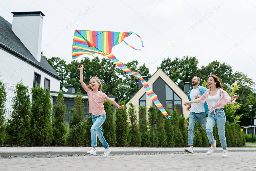 low angle view of happy kid running with colorful kite near cheerful parents on street 