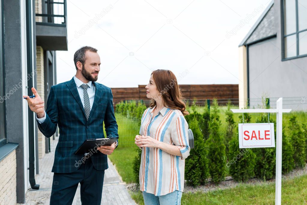 handsome bearded broker holding clipboard and gesturing near attractive woman and board with sale letters 