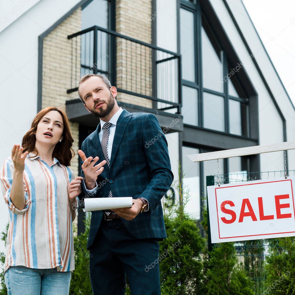 handsome broker holding clipboard and gesturing near beautiful woman and board with sale letters 