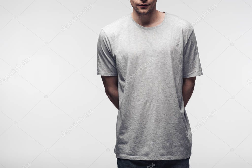 Partial view of man in grey t-shirt holding hands behind back isolated on grey, human emotion and expression concept