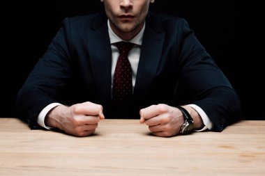 partial view of angry businessman sitting at table and holding fists isolated on black, human emotion and expression concept clipart