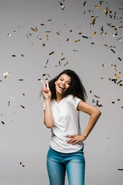 young african american woman smiling near shiny confetti while standing with hand on hip and showing peace sign on grey  clipart