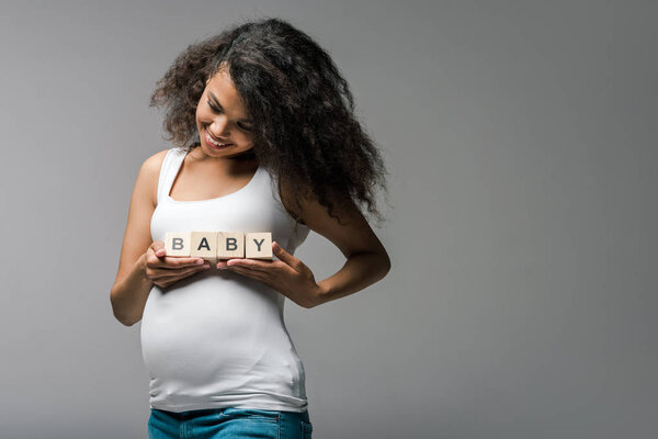 cheerful pregnant african american girl holding wooden cubes with baby lettering on grey 