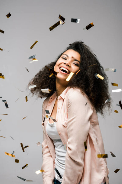 cheerful african american girl smiling near shiny confetti on grey 