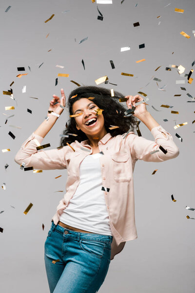 positive young african american woman smiling near shiny confetti while standing on grey 