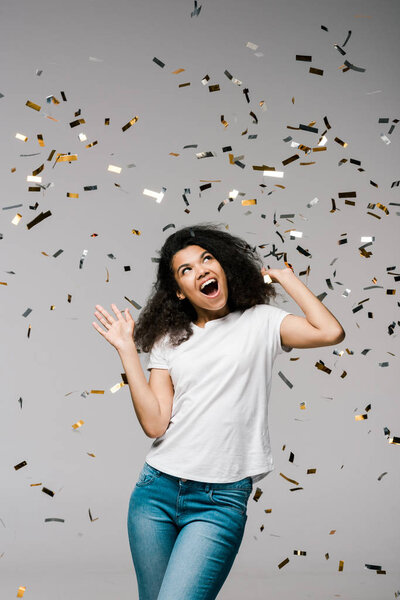 young african american woman smiling and looking at falling confetti while standing on grey 