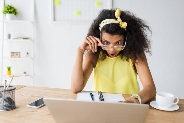 шокированная африканская американка в очках, смотрящая на ноутбук возле чашки кофе
 