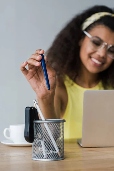 选择性焦点的笔持有人和快乐的非洲裔美国妇女采取笔 而使用笔记本电脑 — 图库照片