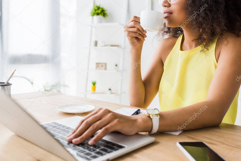 Cropped view of beautiful  african american woman drinking coffee near laptop