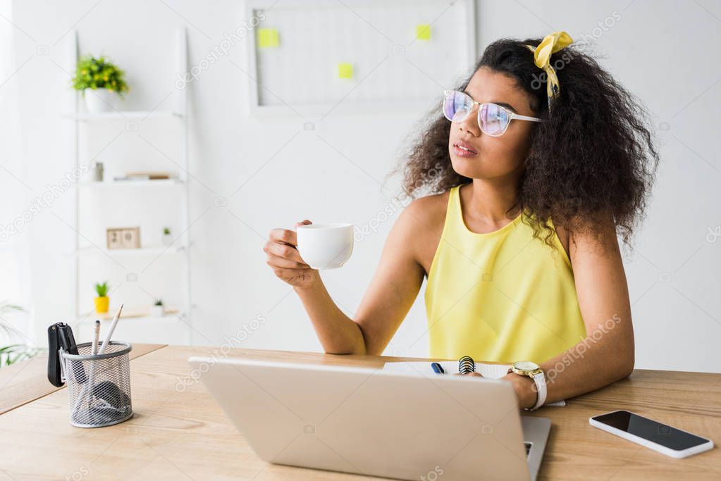 pensive african american girl in glasses holding cup near laptop 