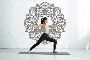 calm asian woman standing with outstretched hands on yoga mat near mandala ornament  clipart