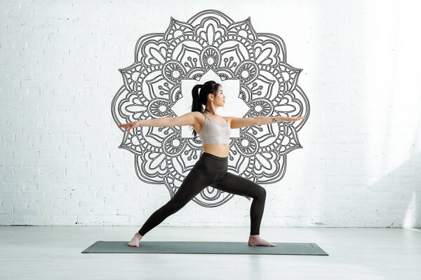 calm asian woman standing with outstretched hands on yoga mat near mandala ornament 