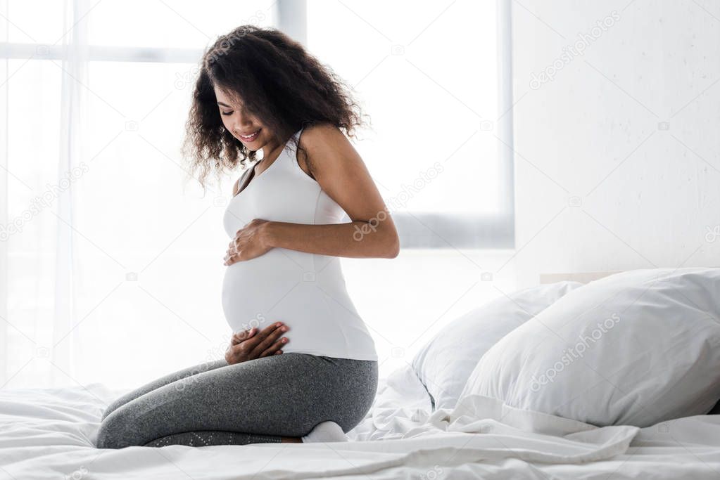 smiling pregnant african american woman touching belly while sitting on bed 