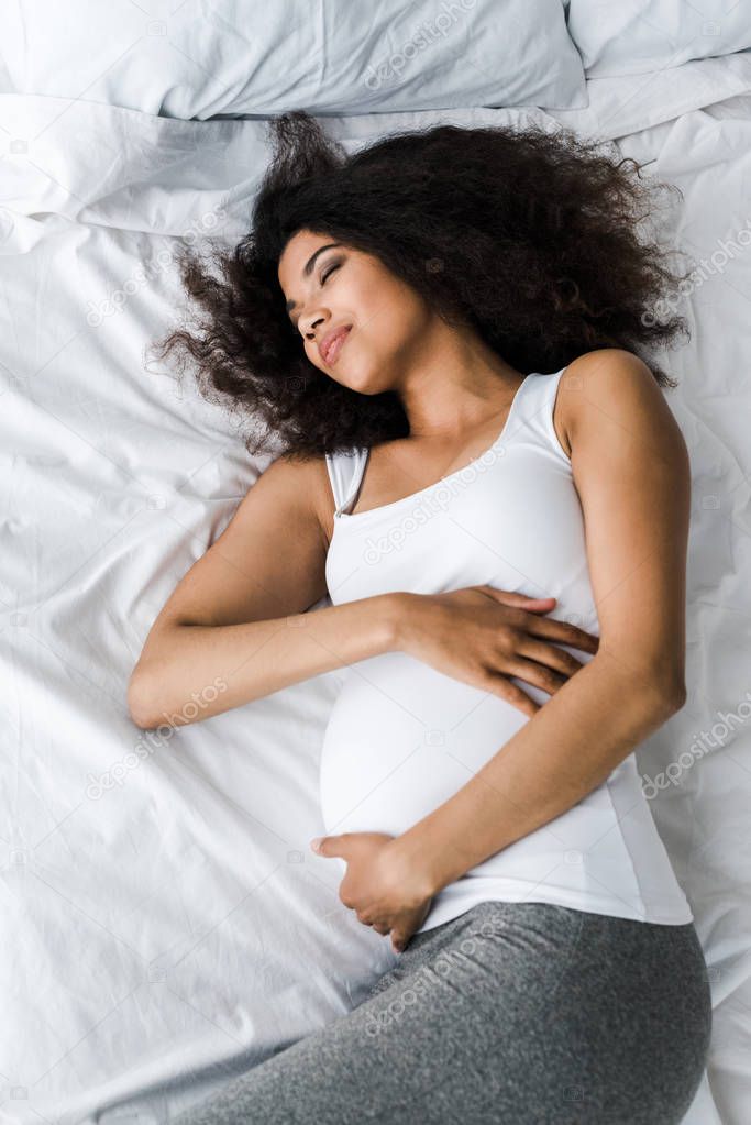 overhead view of happy pregnant african american woman touching belly while lying on bed 