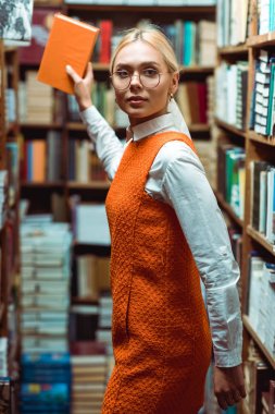 blonde and beautiful woman in glasses and dress holding book and looking at camera in library  clipart