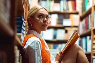 beautiful and blonde woman in glasses holding book and looking away in library  clipart