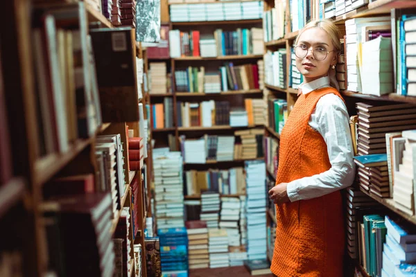 Piękna Blond Kobieta Okularach Pomarańczowa Sukienka Stoi Patrząc Dala Bibliotece — Zdjęcie stockowe