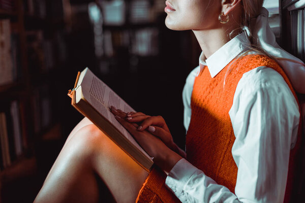 cropped view of woman in orange dress sitting and holding book in library 