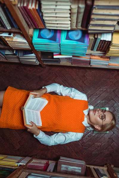 美丽的女人在橙色礼服躺在地板上 拿着书在图书馆的顶视图 — 图库照片