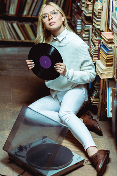 pretty and blonde woman sitting on floor and holding vinyl in library 