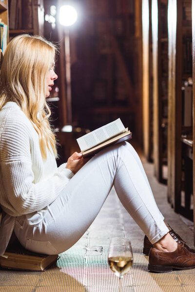 side view of blonde woman reading book and sitting on floor in library 