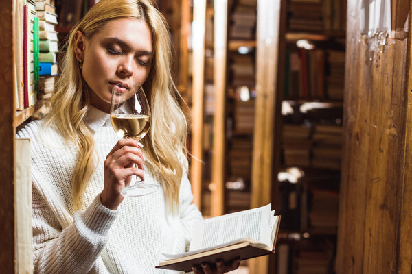 blonde woman with closed eyes holding book and wine glass in library 