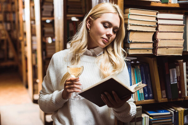 blonde and pretty woman reading book and holding wine glass in library 