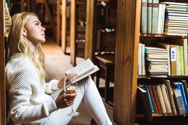 side view of blonde and dreamy woman holding book and wine glass in library 