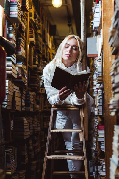 blonde and pretty woman in white sweater reading book in library 