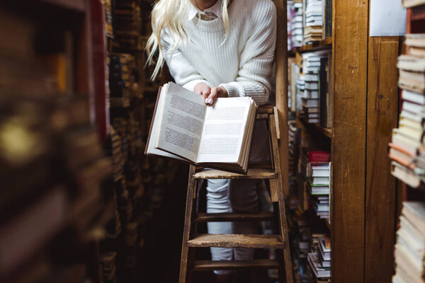 cropped view of woman in white sweater holding book in library 