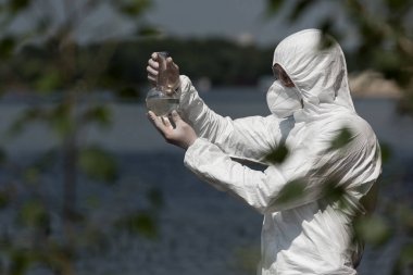 selective focus of water inspector in protective costume, respirator and goggles holding flask with water sample clipart